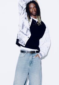 Giacca oversized in bianco e nero con stampa tie-dye, con polsini a coste, indossata sopra un polo nera e una t-shirt bianca; jeans a gamba larga azzurri.