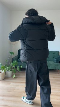 Casaco puffer preto com capuz, apresentando um design acolchoado, ombros largos e um ajuste descontraído. Interior visível perto do fecho.