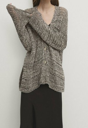 Woman wearing a loose, chunky knit cardigan with large buttons over a black skirt, adjusting the sleeve on a white background.
