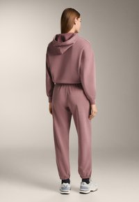 Roze katoenen hoodie met verstelbare trekkoorden en bijpassende joggers. Voorzien van elastische manchetten en tailleband, gecombineerd met grijze sportschoenen.