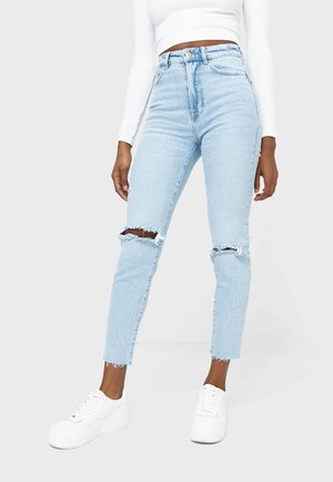 Femme portant un jean skinny bleu clair déchiré, un crop top blanc à manches longues et des baskets à plateforme blanches, debout sur un fond uni.