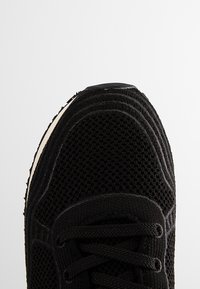 Zbliżenie na czarny siatkowy czubek trampka z sznurowadłami i fakturowanym brzegiem podeszwy na jednolitym białym tle.