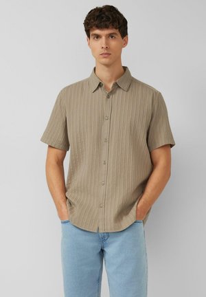 Junger Mann mit kurzen lockigen Haaren trägt ein beigefarbenes kurzärmeliges Hemd mit Knöpfen und hellblaue Jeans, steht mit den Händen in den Taschen.