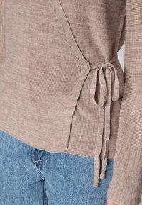 Stickad tröja i mjuk beigefärg med sidband, långa ärmar och ribbad textur. Parat med ljusblå denim, som visar liten nötthet i kanterna.