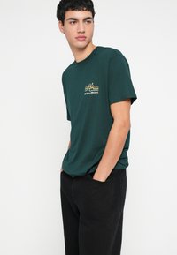 Πράσινο βαμβακερό μπλουζάκι με μικρό χρυσό και πορτοκαλί σχέδιο βουνού στο αριστερό στήθος, συνδυασμένο με μαύρο παντελόνι. Κανονική εφαρμογή και σχεδιασμός με στρογγυλή λαιμόκοψη.