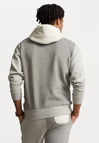 Grå sweatshirt med en vit inre huva, med långa ärmar och en bakficka. Mjuk textur och avslappnad passform.