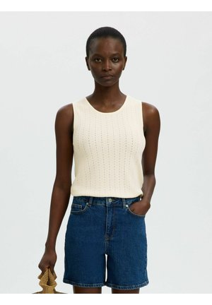 Donna in piedi contro uno sfondo semplice, indossa un top in maglia color crema senza maniche e pantaloncini di jeans blu, una mano in tasca, tiene una borsa.