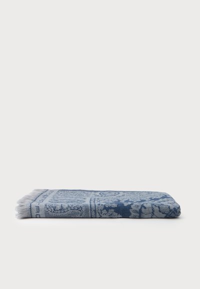Tissu bleu plié avec des motifs circulaires et floraux complexes, orné de franges blanches sur un côté, posé à plat sur une surface blanche.