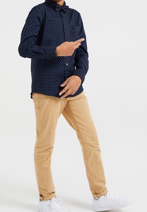 Marineblauses Hemd mit weißen Punkten, knopfbares Design und Fliege; kombiniert mit khakifarbenen Hosen und weißen Sneakers.