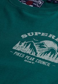 Zielony bawełniany t-shirt z białym graficznym wzorem przedstawiającym góry i drzewa oraz tekstem "SUPER" i "PIKES PEAK COUNCIL", z okrągłym dekoltem.