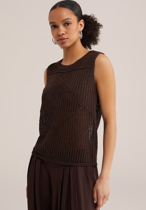 Joven con coleta rizada que lleva pendientes de aro y una camiseta marrón de punto sin mangas con un patrón geométrico, combinada con pantalones marrones sueltos.