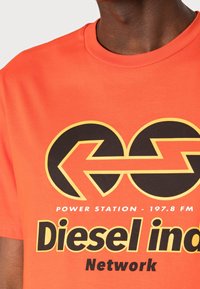 Orange Baumwoll-T-Shirt mit schwarzem und gelbem Grafikdesign, das Pfeile und den Text "Diesel ind. Network" und "Power Station - 197.8 FM" zeigt.