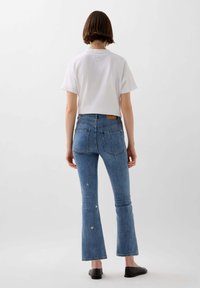 Femme portant une chemise blanche à manches courtes, un jean flare taille haute bleu avec de petites embellissements en argent, et des chaussures noires à enfiler, de dos.