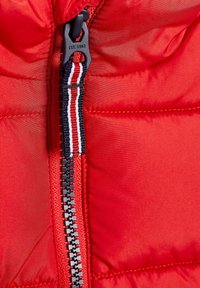 Röd puffjacka med en svart dragkedja och en texturerad dragflik. Synliga sömmar och en trikolor randig detalj nära dragkedjan.