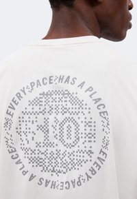 Gros plan d'une personne portant une chemise blanche avec un motif circulaire gris pixellisé et le texte "EVERY:PACE:HAS A PLACE" dans le dos.