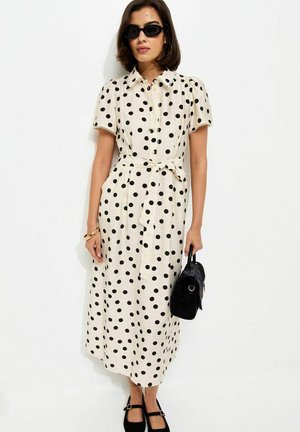 Kobieta w czarnych okularach przeciwsłonecznych i białej sukience midi w czarne groszki, trzymająca czarną torebkę, stojąca na tle białego tła.