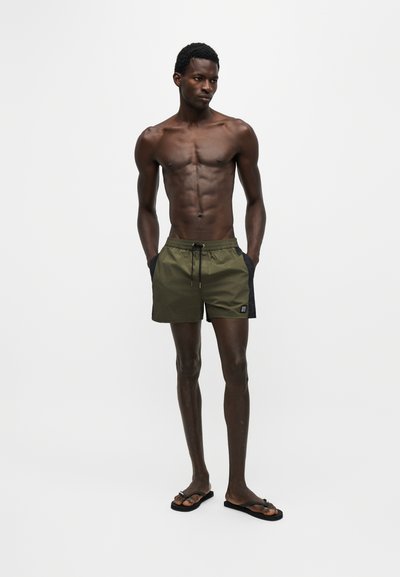 Homme mince à la peau foncée portant un short de bain vert et noir ainsi que des tongs noires, debout les mains dans les poches devant un fond uni.