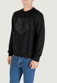 Felpa nera con una grafica testurizzata di una tigre sul davanti, maniche lunghe, polsini a coste e orlo aderente, abbinata a jeans blu.