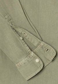 Μανσέτα μακριού μανικιού σε ελαφρύ ελαιώδη πράσινο χρώμα με ένα κουμπί κουμπωμένο και μια κουμπότρυπα ξεκουμπωμένη σε υφασμάτινο φόντο.