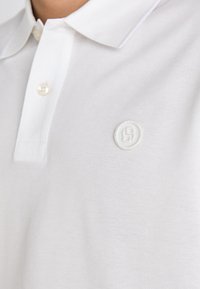 Nærbilde av en hvit pikéskjortekrage med en knapp og et subtilt brodert rundt logo på brystet.