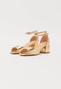 Gouden open-teen hakken met een metallic afwerking, verstelbare enkelriem en een chunky blokhak. Soepele textuur met een minimalistisch ontwerp.