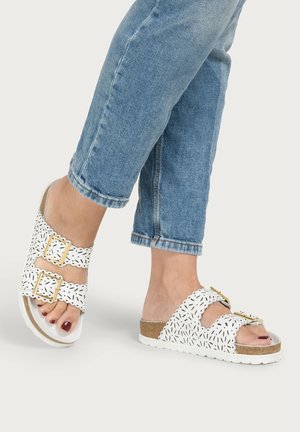 Feet wearing white open-toe sandals with gold buckles and patterned cutouts, paired with light blue rolled-up jeans on a plain background.