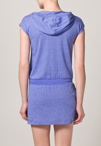 Modrá šatová halenka s kapucí a krátkým rukávem, se zúženým pasem a strukturovanou tkaninou. Délka nad kolena, obsahuje decentní detaily prošití.