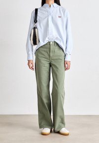 Světle modrá košile na knoflíky s vyšitým logem, v kombinaci s vysokým olivově zeleným širokým kalhotami a bílými teniskami s hnědými podrážkami.