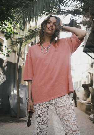 Różowy, luźny t-shirt z krótkimi rękawami i okrągłym dekoltem, zestawiony z białymi koronkowymi spodniami z kwiatowym wzorem. Akcenty w postaci srebrnej biżuterii.