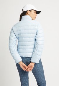 Chaqueta inflable azul claro con diseño acolchado horizontal y cuello alto. La modelo lleva leggings azules con patrón, con las manos unidas por detrás.