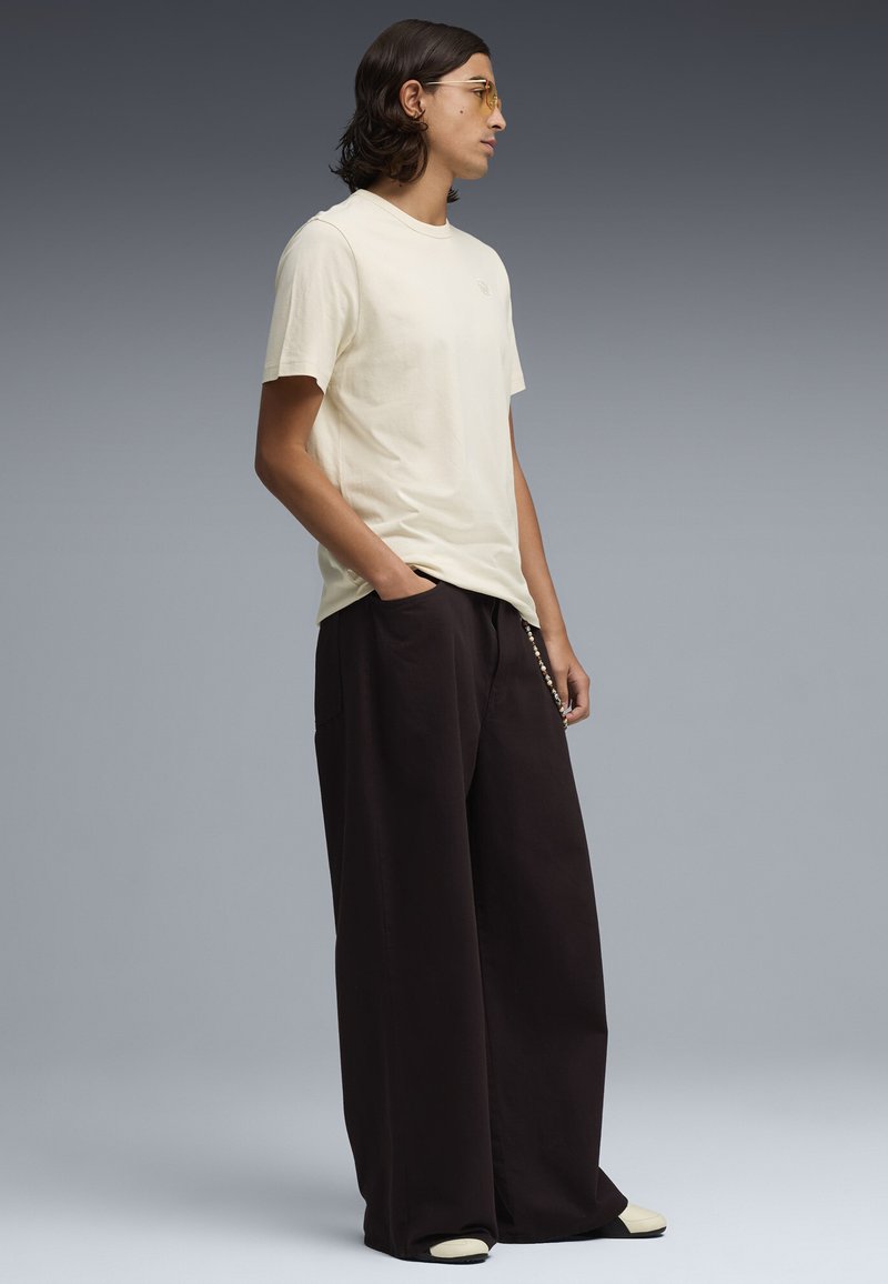 Jeune personne aux cheveux mi-longs portant un t-shirt beige, un pantalon noir surdimensionné et des lunettes teintées jaunes, debout les mains dans les poches.