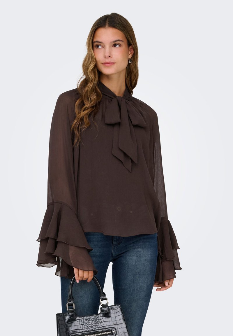 Femme portant un chemisier transparent marron foncé avec un col noué et des manches à volants superposés, un jean bleu, tenant un sac à main noir texturé.