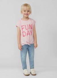 Lichtroze T-shirt met "FUN DAY" in fuzzy roze letters, geknoopt aan de zoom, gecombineerd met lichtblauwe jeans en witte sneakers.
