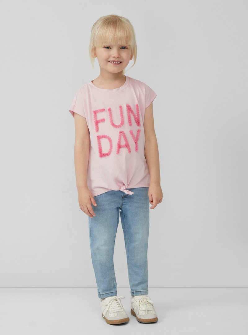 Lichtroze T-shirt met "FUN DAY" in fuzzy roze letters, geknoopt aan de zoom, gecombineerd met lichtblauwe jeans en witte sneakers.