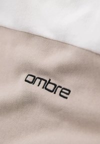 Tejido de algodón en secciones de blanco y beige claro. Presenta texto bordado en negro que dice "ombre" en el área beige. Textura suave, diseño casual.
