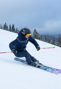 Skifahrende Person in dunkelblauer Jacke und Hose, schwarzem Helm und Skibrille, die pinke und gelbe Skistöcke auf einer verschneiten Piste mit immergrünen Bäumen benutzt.