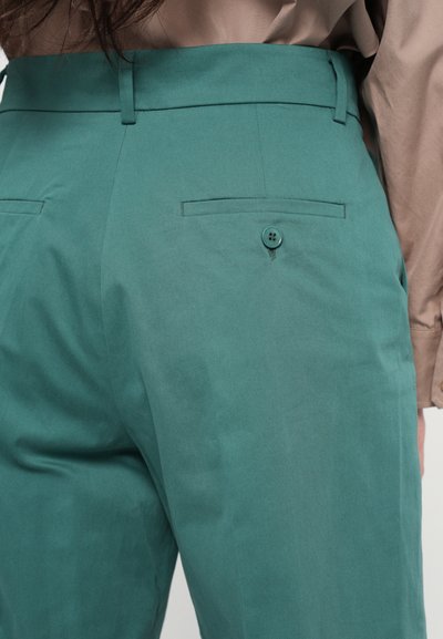 Grüne Hose mit glatter Textur, ausgestattet mit einem einzelnen Knopf und zwei hinteren Taschen. Der Bund ist ordentlich verarbeitet und betont die Passform.