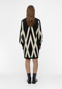 Robe pull tricotée en noir et crème présentant un motif géométrique en zigzag. Manches longues avec bords côtelés, design arrivant au genou.