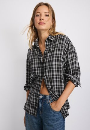 Jeune femme portant une chemise à carreaux noire et blanche avec les manches retroussées et un jean bleu, debout devant un fond clair uni.