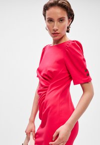 Malinovo ružová šaty s pufovanými rukávmi, drapériou na prednej strane a tromi čiernymi gombíkmi na rukávoch. Hladká tkanina s priliehavým strihom.