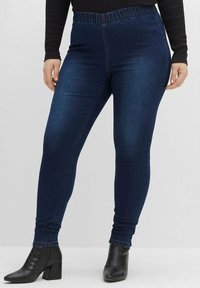 Mörkblå denimleggings med elastisk midja, slim fit och subtila sömdetaljer, kombinerat med svarta klackboots vid vristerna.