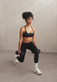 Sujetador deportivo negro con acento rosa, leggings negros con textura lateral, pesas grises y zapatillas deportivas blancas. Posición de zancada sobre un suelo liso.