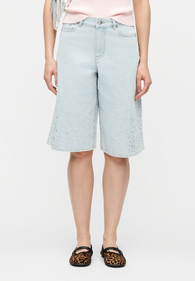 Άτομο που φοράει γαλάζιες τζιν βερμούδες μήκους έως τα γόνατα με μικρές παγιέτες και φλατ παπούτσια με λεοπάρ σχέδιο, στέκεται μπροστά σε απλό φόντο.