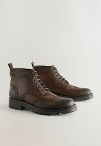 Brune læder ankelstøvler med brogue-detaljer, rund tå og sorte gummisåler. Har snørebåndslukning og strop bagpå.