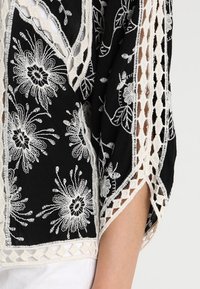 Sort stof top med hvid blomsterbroderi, åbne vævninger og en løs pasform. Har korte ærmer og detaljeret design i nederste kant.