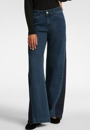 Woman wearing dark blue wide-leg jeans with contrast darker side panels, black top, and black shoes, standing on a light background.