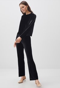 Schwarzes, samtweiches Langarmoberteil, kombiniert mit passenden Hosen. Ausgestellt silhuette mit glatter Textur. Modell trägt beigefarbene Absatzschuhe.