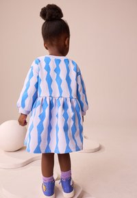 Haljina plave i bijele cik-cak uzorka s dugim rukavima i skupom suknjom, uparena s plavim tenisicama i ljubičastim čarapama. Jednostavan dizajn.