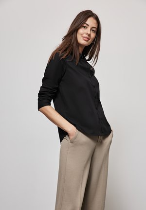 Camicia nera con bottoni, dal tessuto liscio, maniche lunghe e una vestibilità rilassata, abbinata a pantaloni beige a gamba larga.