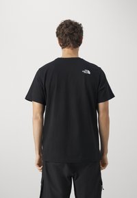 Mann mit lockigem Haar, der ein schwarzes The North Face T-Shirt und schwarze Hosen trägt, von hinten vor einem schlichten Hintergrund betrachtet.